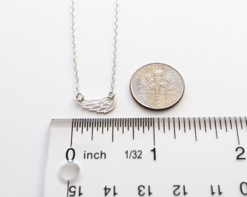 May include: A silver-toned necklace featuring a small wing-shaped pendant. The pendant is positioned next to a United States dime and a clear plastic ruler, showing the wing charm measures approximately 0.75 inches in total width.