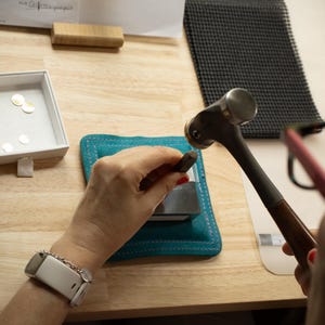 K&ouml;nnte beinhalten: Ein Schmuckhersteller verwendet Hammer und Stempel auf einem Metallst&uuml;ck, mit einer t&uuml;rkisfarbenen Matte und einer Schale mit goldenen Scheiben in der N&auml;he. Die Szene spielt sich auf einer h&ouml;lzernen Arbeitsfl&auml;che ab.
