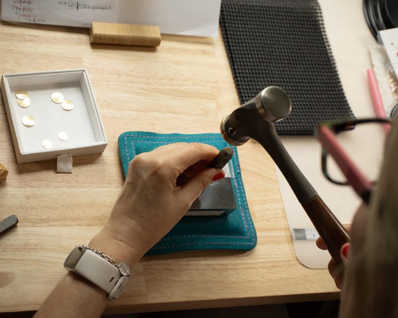 K&ouml;nnte beinhalten: Ein Schmuckhersteller verwendet Hammer und Stempel auf einem Metallst&uuml;ck, mit einer t&uuml;rkisfarbenen Matte und einer Schale mit goldenen Scheiben in der N&auml;he. Die Szene spielt sich auf einer h&ouml;lzernen Arbeitsfl&auml;che ab.