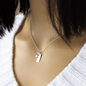 K&ouml;nnte beinhalten: Eine silberne Halskette mit einem rechteckigen Anh&auml;nger und einem kleinen Charm. Der Anh&auml;nger hat ge&auml;tzte Details und h&auml;ngt an einer zarten Kette. Die Halskette wird &uuml;ber einem wei&szlig;en Pullover getragen.