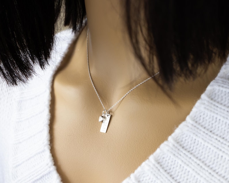 K&ouml;nnte beinhalten: Eine silberne Halskette mit einem rechteckigen Anh&auml;nger und einem kleinen Charm. Der Anh&auml;nger hat ge&auml;tzte Details und h&auml;ngt an einer zarten Kette. Die Halskette wird &uuml;ber einem wei&szlig;en Pullover getragen.