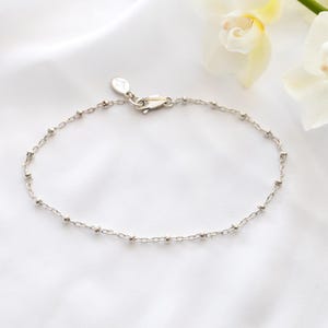May include: A delicate silver bracelet with small, evenly spaced beads and a lobster clasp. The bracelet is displayed on a white, textured surface, with white and yellow orchids in the background.