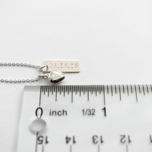 K&ouml;nnte beinhalten: Eine silberne Halskette mit einem kleinen Herzanh&auml;nger und einem rechteckigen Anh&auml;nger mit dem eingravierten Datum "05.21.25". Die Halskette wird neben einem Lineal gezeigt, der Anh&auml;nger misst ca. 0,6 cm.