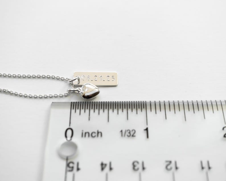 K&ouml;nnte beinhalten: Eine silberne Halskette mit einem kleinen Herzanh&auml;nger und einem rechteckigen Anh&auml;nger mit dem eingravierten Datum "05.21.25". Die Halskette wird neben einem Lineal gezeigt, der Anh&auml;nger misst ca. 0,6 cm.