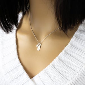 K&ouml;nnte beinhalten: Eine silberne Halskette mit einem kleinen rechteckigen Anh&auml;nger und einem Charm. Der Anh&auml;nger hat eine strukturierte Oberfl&auml;che und eine kleine eingravierte Zahl 1. Der Charm ist ein kleiner Stein. Die Halskette wird &uuml;ber einem wei&szlig;en Pullover getragen.