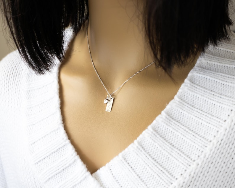 K&ouml;nnte beinhalten: Eine silberne Halskette mit einem kleinen rechteckigen Anh&auml;nger und einem Charm. Der Anh&auml;nger hat eine strukturierte Oberfl&auml;che und eine kleine eingravierte Zahl 1. Der Charm ist ein kleiner Stein. Die Halskette wird &uuml;ber einem wei&szlig;en Pullover getragen.