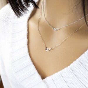 May include: A layered necklace set featuring two delicate chains, each adorned with a small, open-work metal wing charm. One chain is silver-toned and the other is gold-toned, both resting against a white ribbed knit garment.