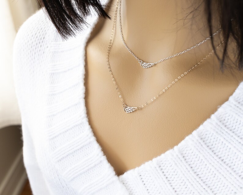May include: A layered necklace set featuring two delicate chains, each adorned with a small, open-work metal wing charm. One chain is silver-toned and the other is gold-toned, both resting against a white ribbed knit garment.