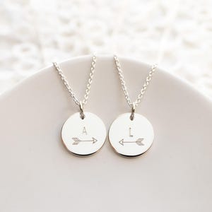 May include: Two silver necklaces with round pendants featuring the letters "A" and "I" with arrows pointing in opposite directions.