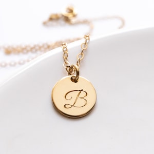 May include: A gold-colored necklace with a circular pendant. The pendant features the cursive letter "B" engraved in the center. The necklace chain and pendant are displayed against a white background.