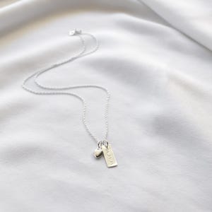 K&ouml;nnte beinhalten: Eine zarte Silberkette mit einem kleinen Herzanh&auml;nger und einem rechteckigen Anh&auml;nger mit Text. Die Kette wird auf einem wei&szlig;en, strukturierten Stoffhintergrund pr&auml;sentiert.