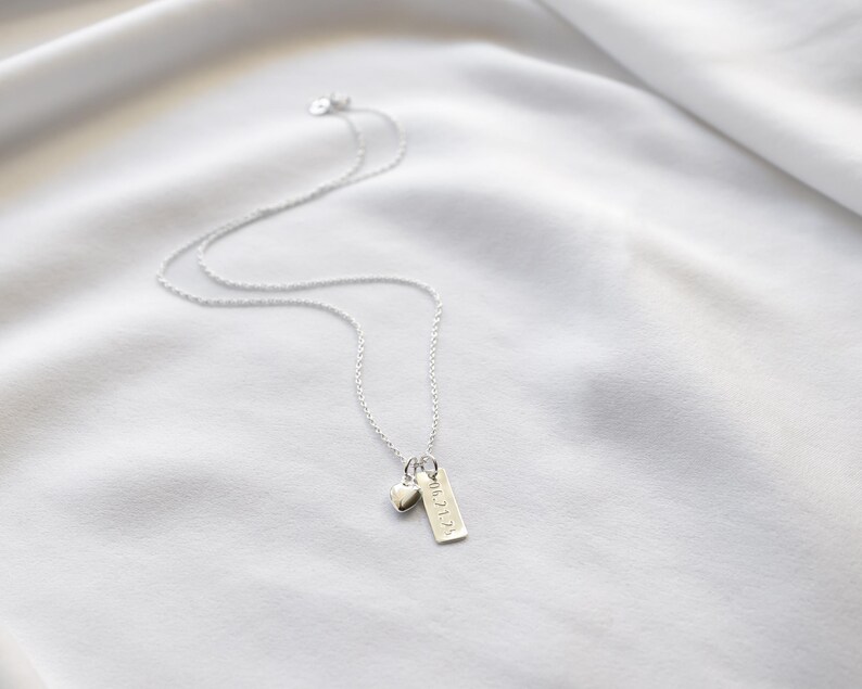 K&ouml;nnte beinhalten: Eine zarte Silberkette mit einem kleinen Herzanh&auml;nger und einem rechteckigen Anh&auml;nger mit Text. Die Kette wird auf einem wei&szlig;en, strukturierten Stoffhintergrund pr&auml;sentiert.