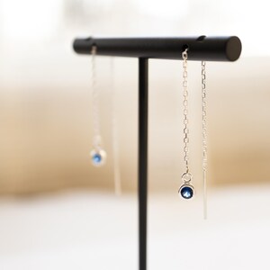 May include: A pair of silver earrings with a small blue gemstone dangling from each earring. The earrings are hanging on a black stand.
