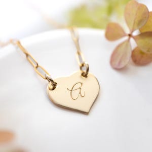 May include: A gold-colored necklace with a heart-shaped pendant. The pendant has a cursive letter "a" engraved on it. The necklace chain is made of rectangular links. The background is a white surface with blurred flowers.