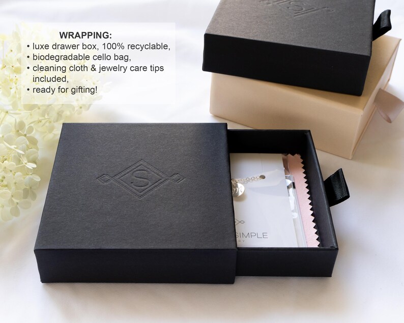 May include: A black square drawer box with a debossed letter S logo on the lid, partially open to reveal a white jewelry card and a pink cleaning cloth inside. A second beige drawer box is stacked behind it, both featuring black pull tabs.