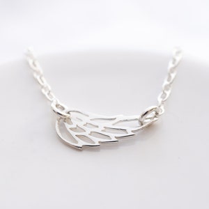May include: A silver-toned necklace featuring a delicate, openwork wing pendant. The pendant is attached to a fine, polished silver-toned chain that extends from both sides of the wing, creating a simple and clean design for this piece of jewelry.