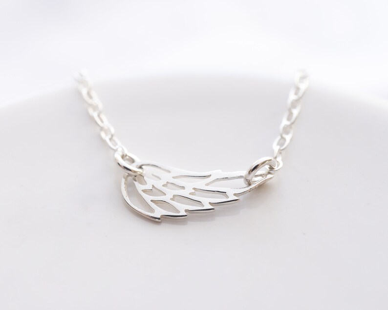 May include: A silver-toned necklace featuring a delicate, openwork wing pendant. The pendant is attached to a fine, polished silver-toned chain that extends from both sides of the wing, creating a simple and clean design for this piece of jewelry.