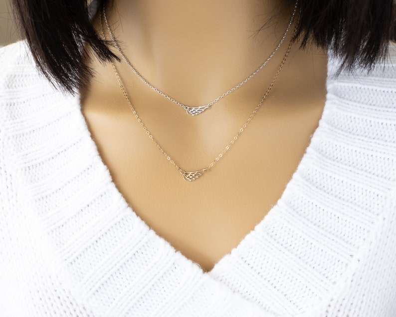 May include: A layered necklace set featuring two delicate chains. The top chain is silver-toned with a small wing-shaped pendant, while the bottom chain is gold-toned and also features a small wing-shaped pendant. Both chains have a fine link design.