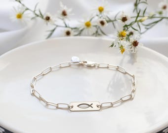 May include: A silver-toned chain bracelet featuring a rectangular metal charm with a cutout fish symbol. The bracelet is constructed with elongated oval links and includes a small circular tag near the lobster claw clasp closure.