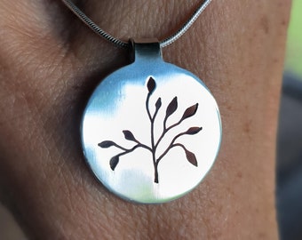 May include: A silver necklace with a round pendant featuring a cut-out design of a tree branch with leaves.