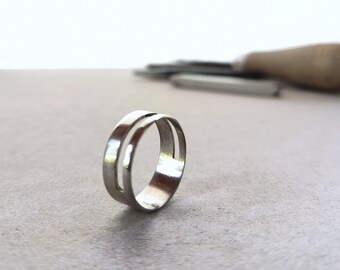 Könnte beinhalten: Ein silberner Ring mit einem einzigartigen Design. Der Ring hat ein Doppelband mit einem rechteckigen Ausschnitt. Der Ring liegt auf einer hellgrauen Oberfläche, mit Werkzeugen im Hintergrund.