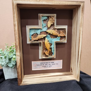 May include: A wooden framed cross with a blue and brown background. The cross is decorated with four fish in a 3D design. The quote "AND HE SAID TO THEM, "FOLLOW ME, AND I WILL MAKE YOU FISHERS OF MEN." Matthew 4:19" is printed on a white tag below the cross.
