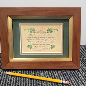 May include: A framed print with a green background and a white mat. The print features a handwritten poem in calligraphy with a green shamrock design. The poem reads: "May the road rise to meet you, May the wind be always at your back, May the sun shine warm upon your face, The rains fall soft upon your fields, And until we meet again, May God hold you, In the palm of His hand."