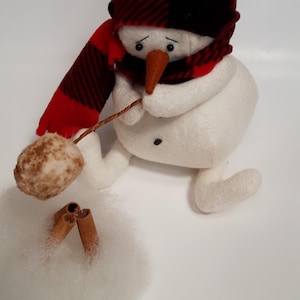 May include: A whimsical snowman figure made of white felt, wearing a red and black plaid hat and scarf. It holds a brown ball on a stick and is next to cinnamon sticks and faux snow. A charming winter decoration.