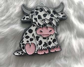 May include: A black and white Highland cow ornament with pink accents. The cow is sitting down with its head tilted to the side. The ornament is made of a textured material and has a rustic look.