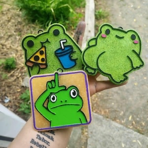 May include: Three flat, decorative frog-themed ornaments. One frog holds a slice of pizza and a blue drink, another is a plain green frog with pink cheeks, and the third is a square piece featuring a green frog making an L-shape with its hand.