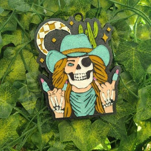 May include: A black and white illustration of a skull wearing a turquoise cowboy hat and bandana, giving the rock and roll hand gesture. The background is black with a crescent moon and cactus.