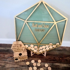 May include: A wooden hexagonal guest book frame with a gold trim and a green background. The frame is standing on a wooden stand. The frame has a sign that says "Welcome, please sign a hexagon and drop it into our frame so we will never forget our loved ones that came". The frame also has a sign that says "Matt and Sara 11.01.2024".