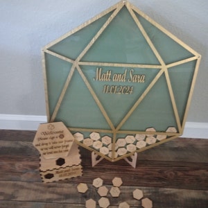 May include: A wooden hexagonal frame with a green glass panel and gold trim. The frame is filled with small wooden hexagonal pieces that guests can sign. The frame is decorated with the names "Matt and Sara" and the date "11.01.2024".