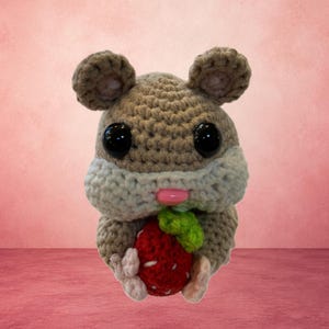 May include: A crocheted hamster toy made with light brown and cream yarn, featuring large black plastic eyes and a small pink plastic nose. The hamster is holding a red crocheted strawberry with a green leaf in its small front paws.