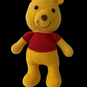 May include: A crocheted Winnie the Pooh bear plush toy with a red shirt. The bear has a yellow body, brown nose, and black eyes.