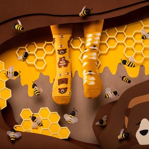May include: A pair of yellow socks with a brown bear and honeycomb pattern. The socks are surrounded by honeycombs and bees.