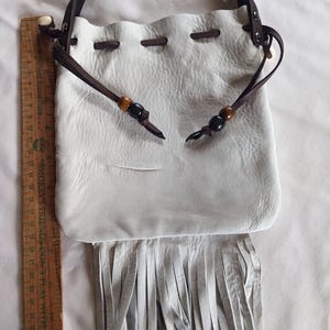 May include: White leather drawstring bag with a brown leather strap and fringe. The bag has wooden and black beads on the strap.