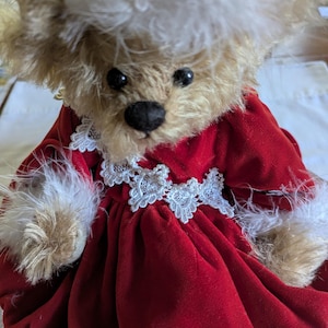 May include: A plush teddy bear wearing a red velvet dress with white lace trim and feather accents. The bear has a light brown fur coat, black eyes, and a black nose. The bear's head is adorned with a white feathered hat.