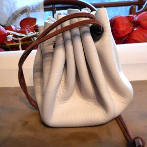 May include: A white leather drawstring pouch with a brown leather cord and a brown leather toggle. The pouch is empty and has a slightly wrinkled appearance.