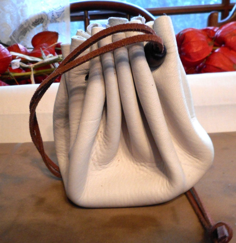 May include: A white leather drawstring pouch with a brown leather cord and a brown leather toggle. The pouch is empty and has a slightly wrinkled appearance.
