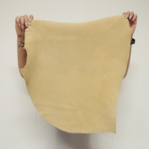 May include: A piece of tan-colored suede leather is held up against a white background. The leather has a soft, textured surface and is being held by two hands, showcasing its shape and size.