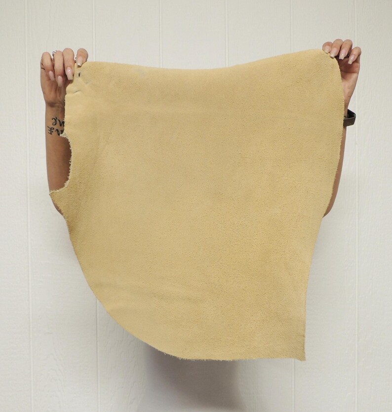 May include: A piece of tan-colored suede leather is held up against a white background. The leather has a soft, textured surface and is being held by two hands, showcasing its shape and size.