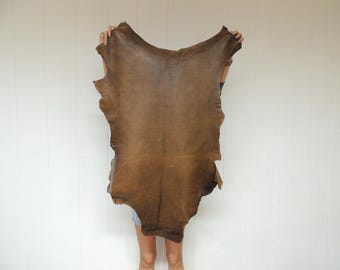 May include: A large, irregularly shaped piece of brown leather is held up against a white background. The leather has a textured surface and varying shades of brown, with darker areas along the edges. The material appears to be tanned and ready for use.