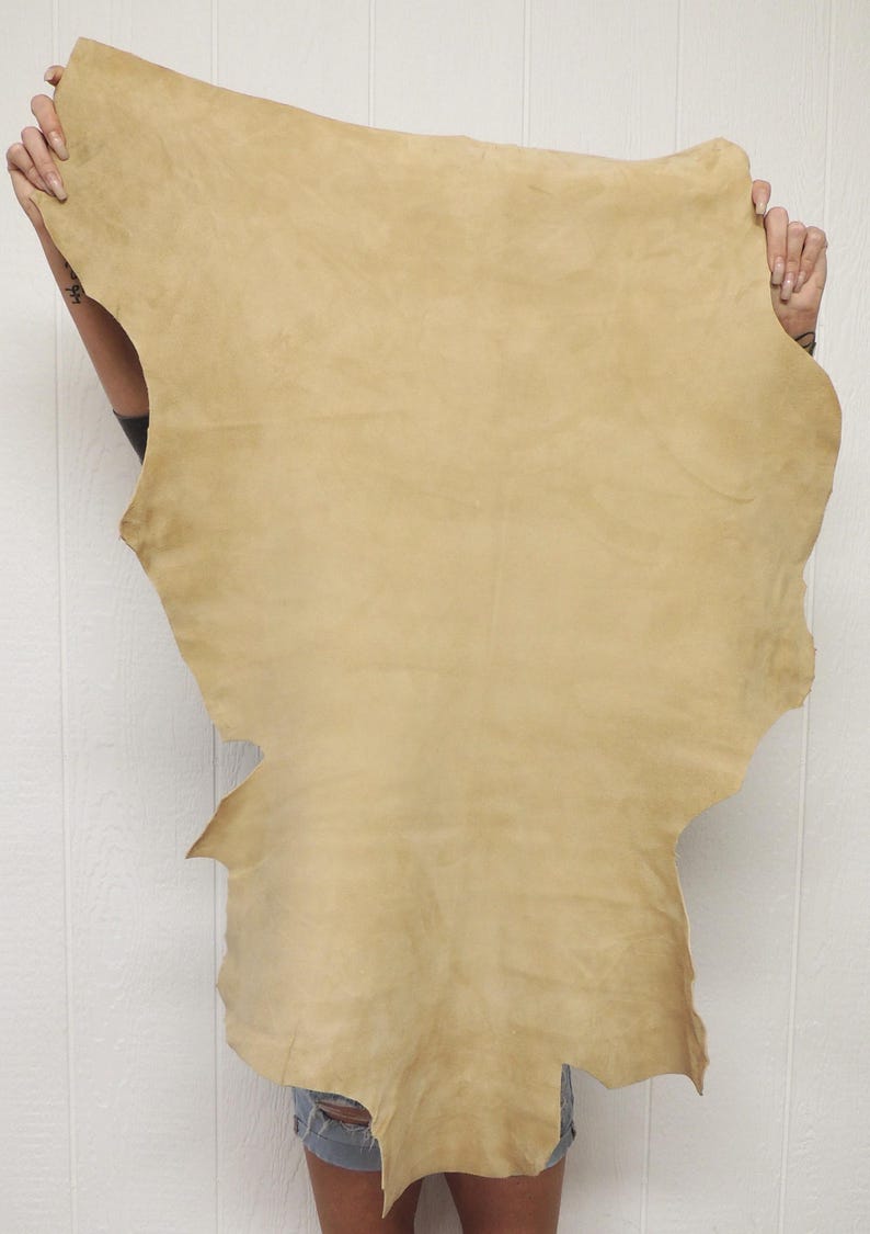 May include: A large, irregularly shaped piece of tan-colored leather. The leather has a soft, suede-like texture and is held up against a white background. The edges of the leather are uneven, suggesting it is a natural hide.
