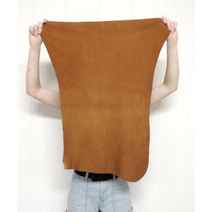 May include: A large, rectangular piece of brown leather is held up by a person. The leather has a smooth texture and a warm, earthy tone. The edges are slightly curved, and the material appears to be flexible.