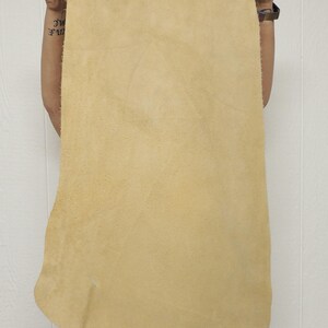 May include: A large, rectangular piece of tan-colored leather. The leather has a textured surface and is held up by a person. The edges of the leather are uneven. The background is a white wall.