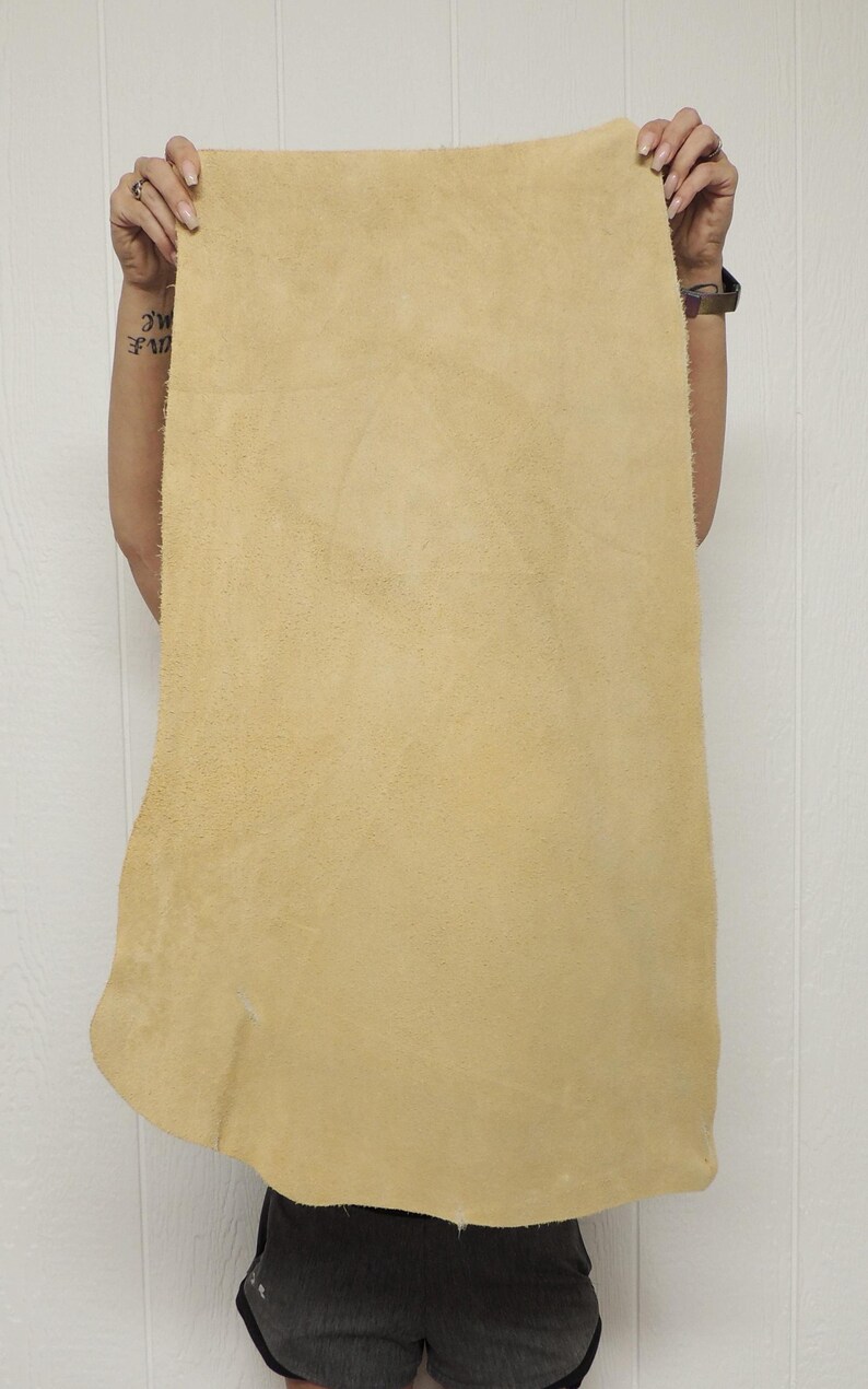 May include: A large, rectangular piece of tan-colored leather. The leather has a textured surface and is held up by a person. The edges of the leather are uneven. The background is a white wall.