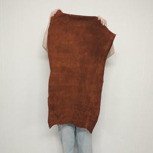 May include: A large, irregularly shaped piece of brown leather is held up against a white background. The leather has a textured surface and a rich, warm color, with variations in tone. The edges are uneven, suggesting a natural, unfinished state.
