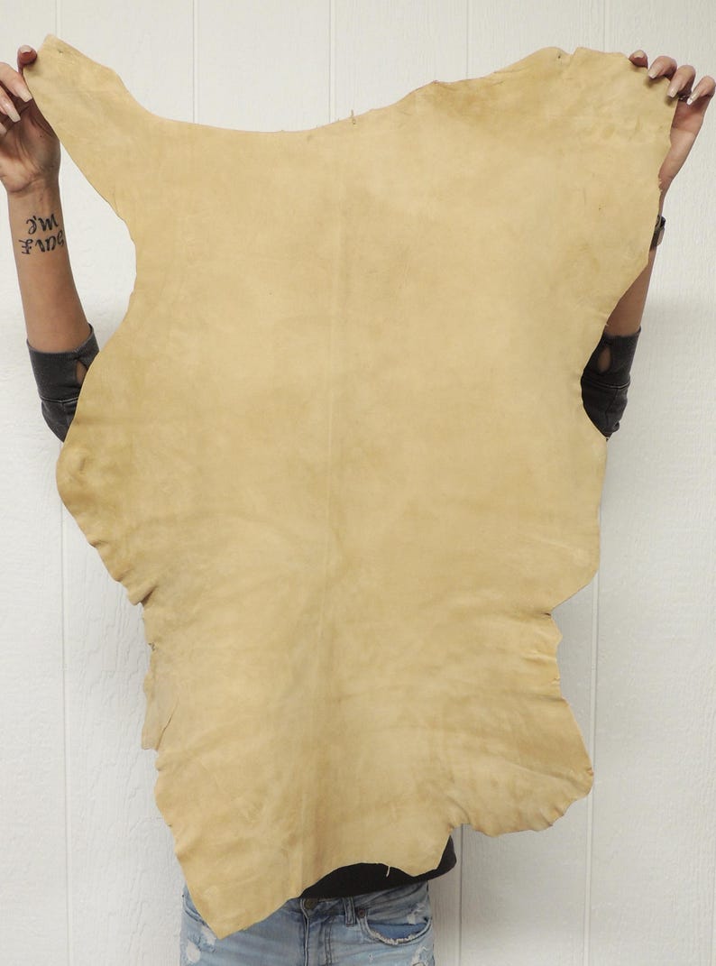 May include: A large, irregularly shaped piece of tan leather. The leather is held up, showing its natural edges and texture. The color is a warm, neutral tone, suitable for various crafting projects.