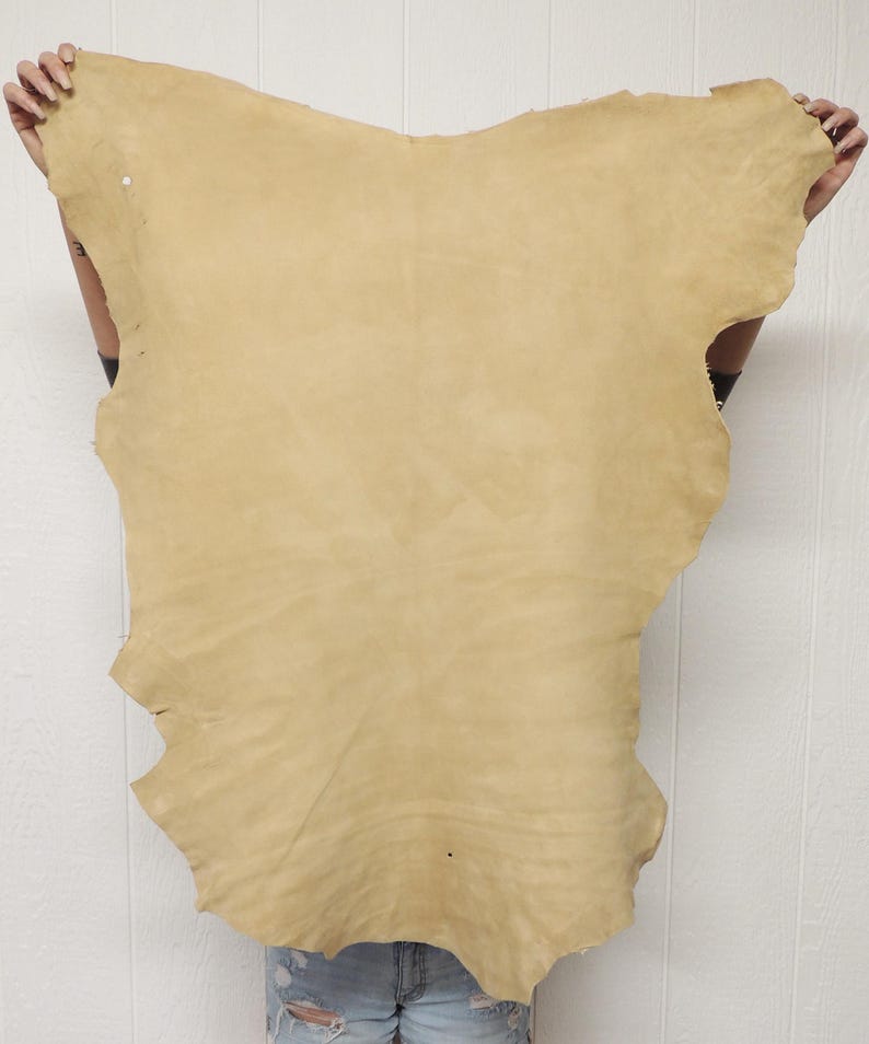 May include: A large, irregularly shaped piece of tan leather. The leather has a natural, uneven edge and a slightly textured surface. The hide is held up against a white background, showcasing its color and form. This leather is suitable for crafting.